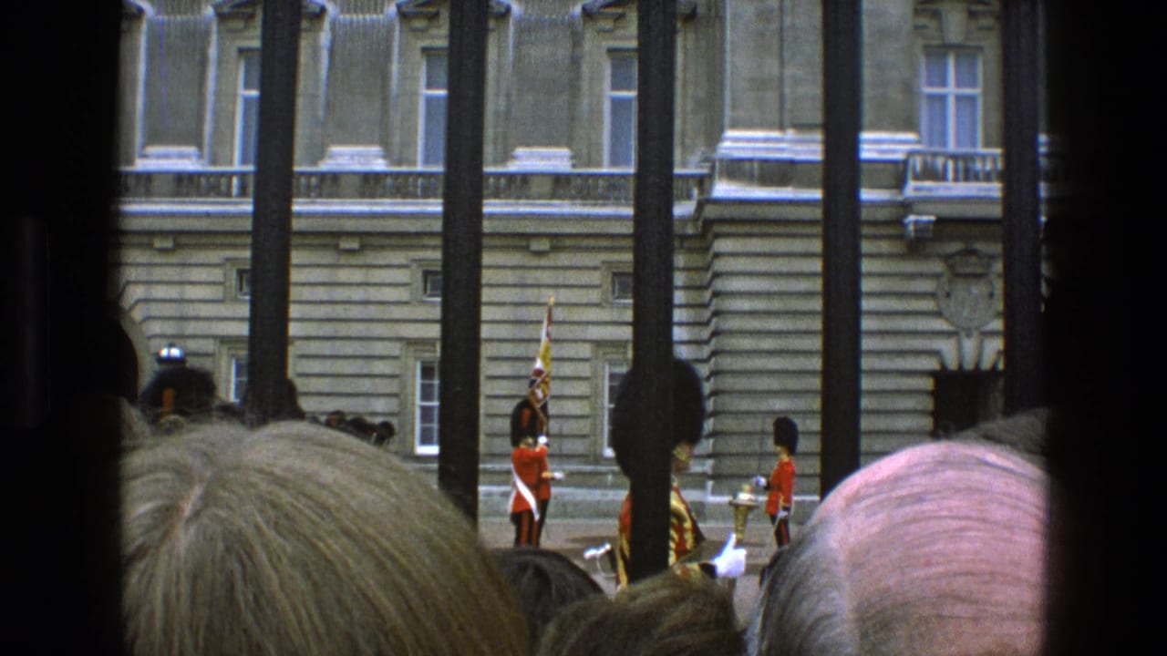 1969 England home-movie view of onlookers watching red-coated royal guards through dark vertical bars before a stone palace facade.