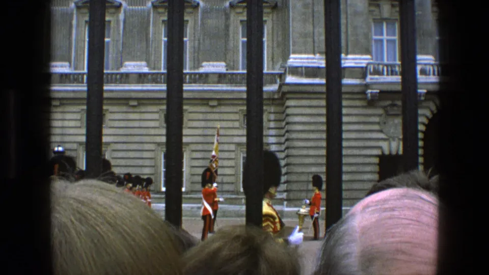 A crowd looks through iron bars toward red-uniformed guards standing before a large stone building in England, 1969.