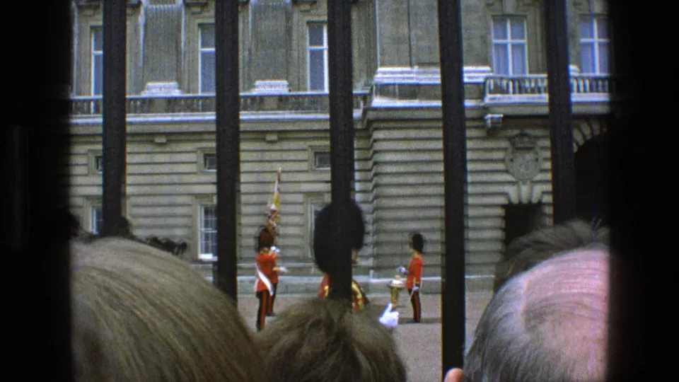 Obstructed 8mm view of royal guards in red tunics behind dark railings, with spectators' heads in the lower foreground.