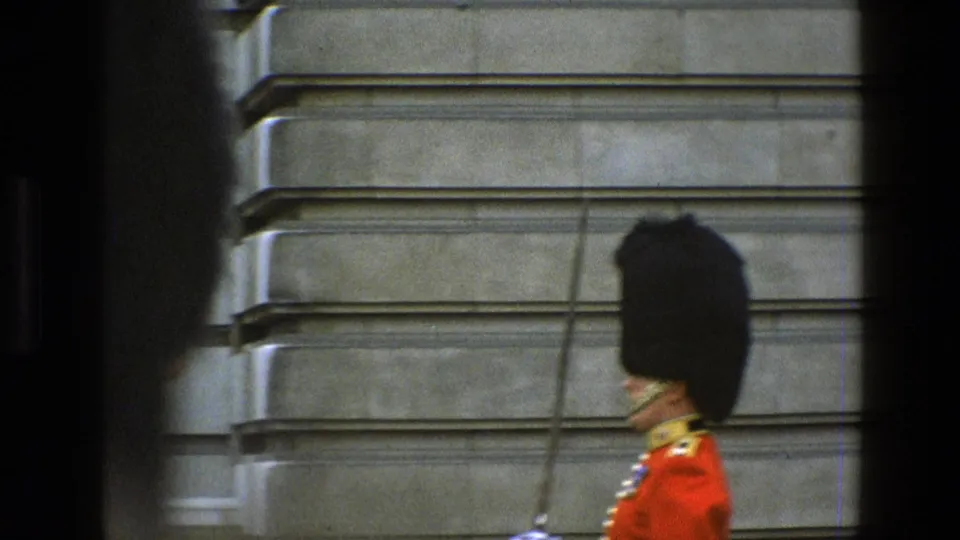 Closer side view of a red-coated guard in a tall black hat beside a stone palace wall, filmed in England in 1969.