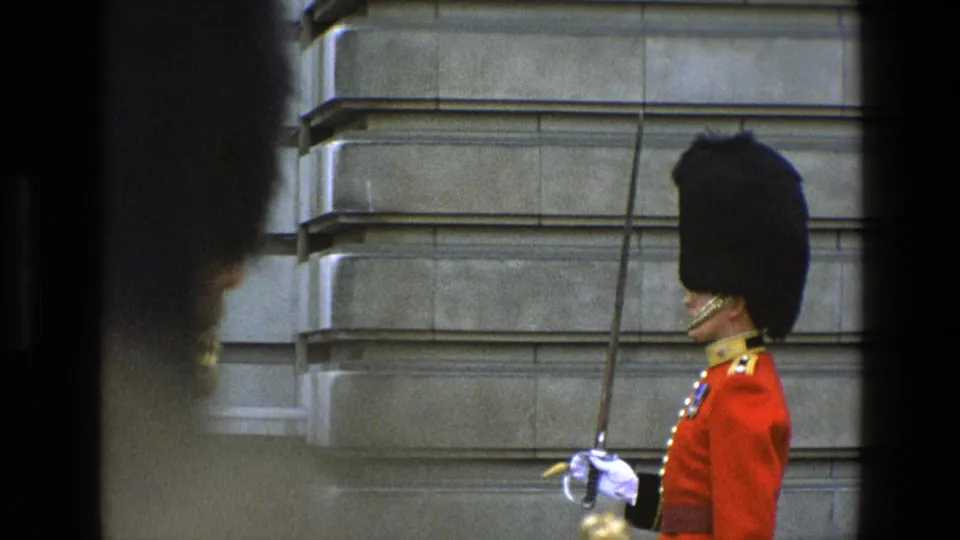 1969 England frame of a uniformed ceremonial guard in profile, wearing a tall black headdress and holding a sword before stone masonry.