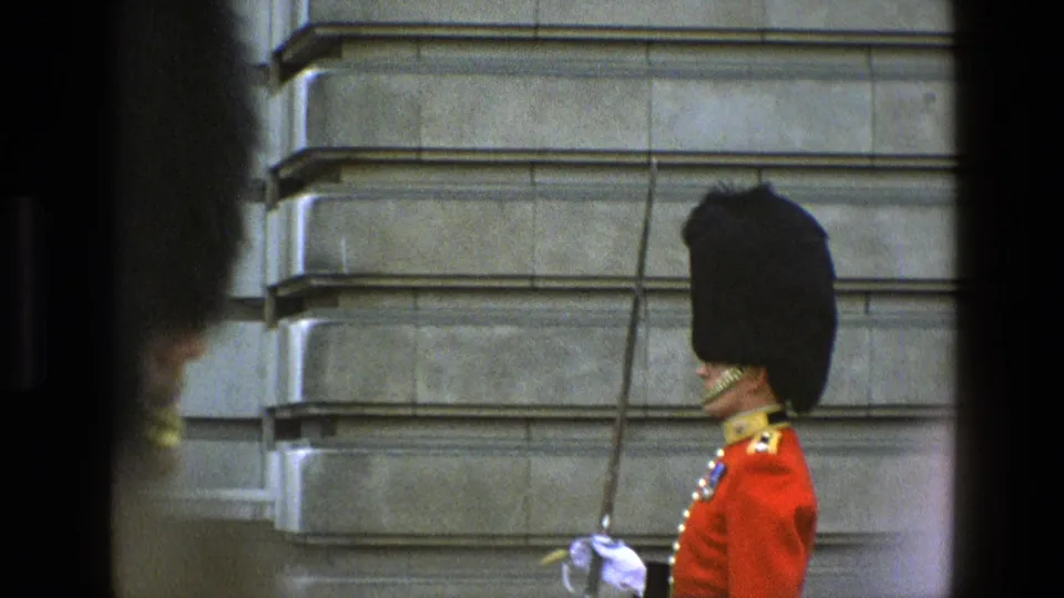 1969 England home-movie frame showing a ceremonial guard in red uniform and black headdress, partly obscured by foreground shadows.