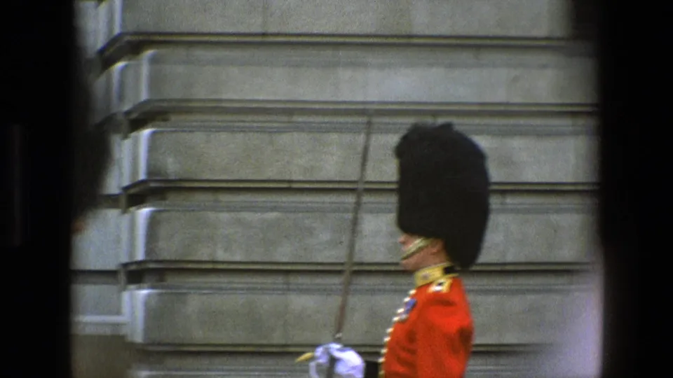 1969 England frame with a ceremonial guard in red tunic, black headdress, and white gloves holding a sword beside a stone wall.