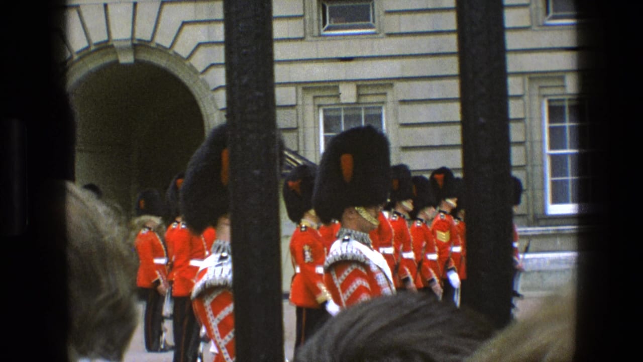 Grenadier Guards in red tunics stand outside a stone palace facade behind dark vertical bars.