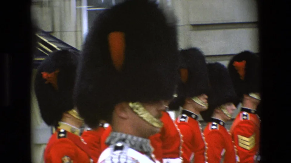 Close 8mm frame of ceremonial guards outside Buckingham Palace in 1969, with faces partly shaded by bearskin hats and foreground bars.