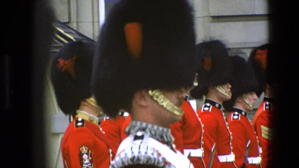 1969 England 8mm frame showing ceremonial guards in red tunics and tall black hats, partly obscured by a foreground guard and dark framing.