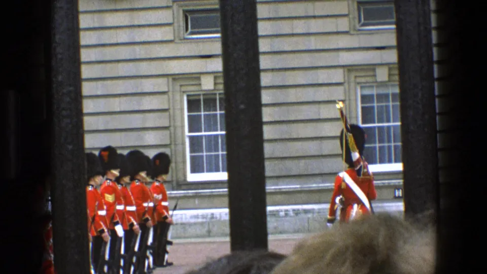 1969 England 8mm frame showing a line of ceremonial guards and a color bearer outside a stone building, filmed through vertical bars.