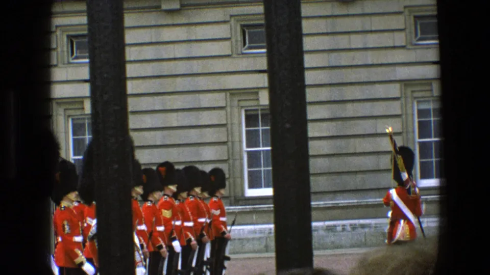 1969 England home-movie frame of ceremonial guards standing outside a stone building, with dark bars dividing the spectator's view.