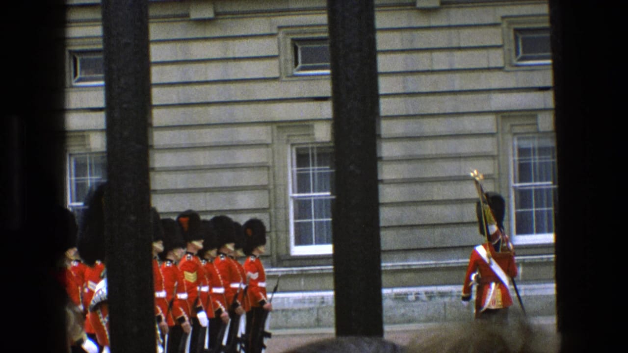 In 1969 England, red-uniformed ceremonial guards with black hats stand in formation outside a formal stone facade, viewed through foreground bars.
