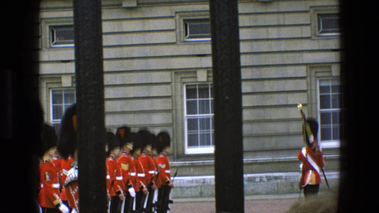 A line of red-coated guards stands near a stone building while a guard with a raised rifle stands apart.