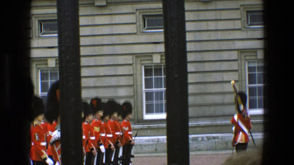 In 1969 England, a ceremonial guard formation stands beside a grand stone building, with one guard holding a rifle upright at the edge of the frame.