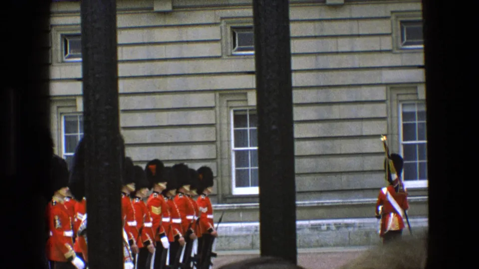In 1969 England, a visitor-shot view through dark bars shows ceremonial guards in red tunics and black hats aligned outside a stone building.