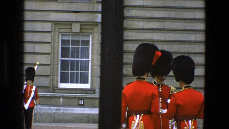 In 1969 England, three ceremonial guards in red tunics and tall black hats are filmed closer near a stone building window, with one face partly visible.