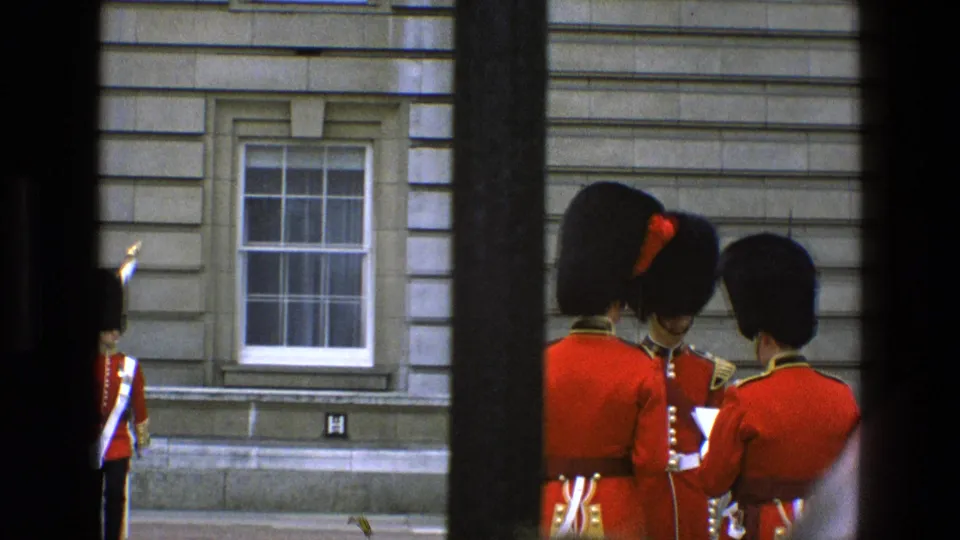 Three royal guards in red tunics and black bearskin hats appear to converse outside Buckingham Palace in 1969 footage.