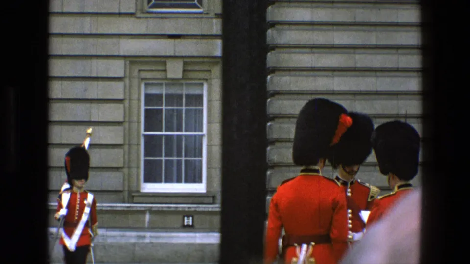 Royal guards in red tunics stand near Buckingham Palace as another guard with ceremonial gear appears in the background.