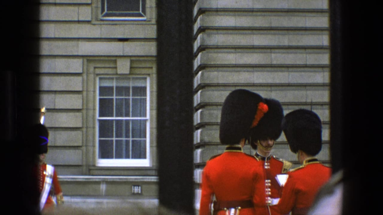 1969 England 8mm frame of ceremonial guards in red tunics and bearskin hats gathered near a stone building.
