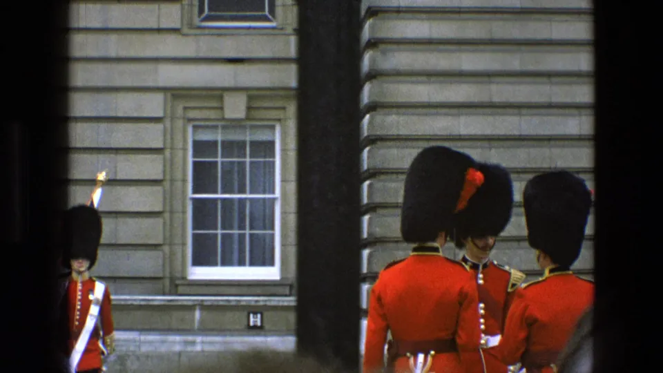 1969 England 8mm frame showing several ceremonial guards by a stone building, partly framed by dark vertical bars.
