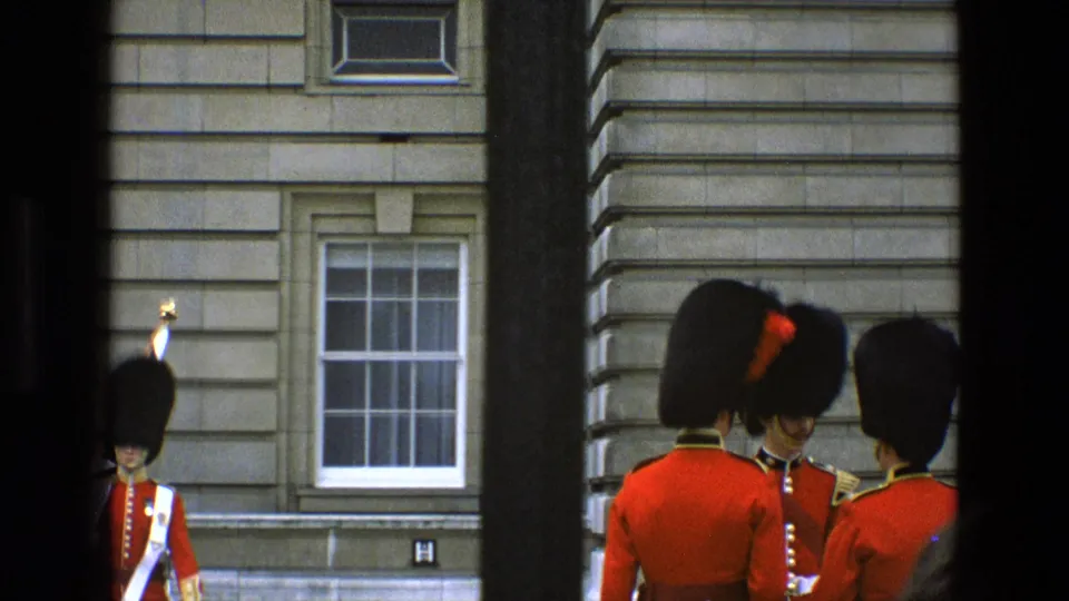 1969 England 8mm frame of red-coated ceremonial guards standing close together near a stone facade and sash window.