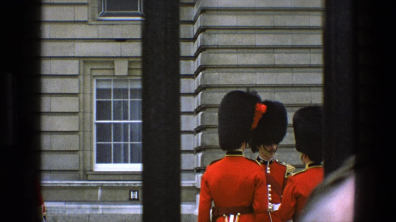 British guards in red uniforms stand behind dark vertical railings outside a stone building.
