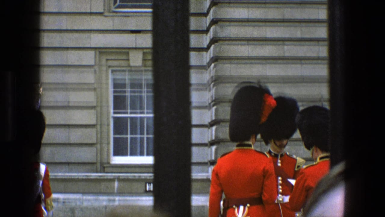 British ceremonial guards outside Buckingham Palace appear in conversation, partly obscured by dark railings in a 1969 home-movie frame.