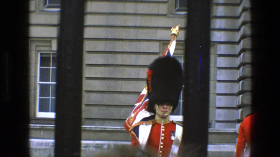 British guard in red ceremonial dress is framed by dark vertical bars against Buckingham Palace stonework in 1969 footage.
