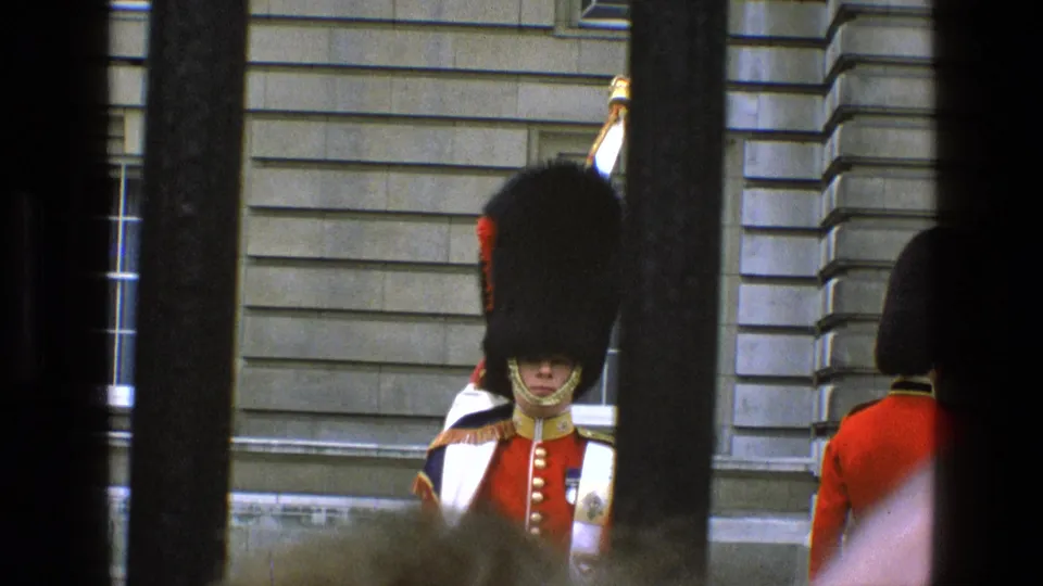 1969 England 8mm frame showing a royal guard in red tunic and bearskin hat viewed through dark gate bars.