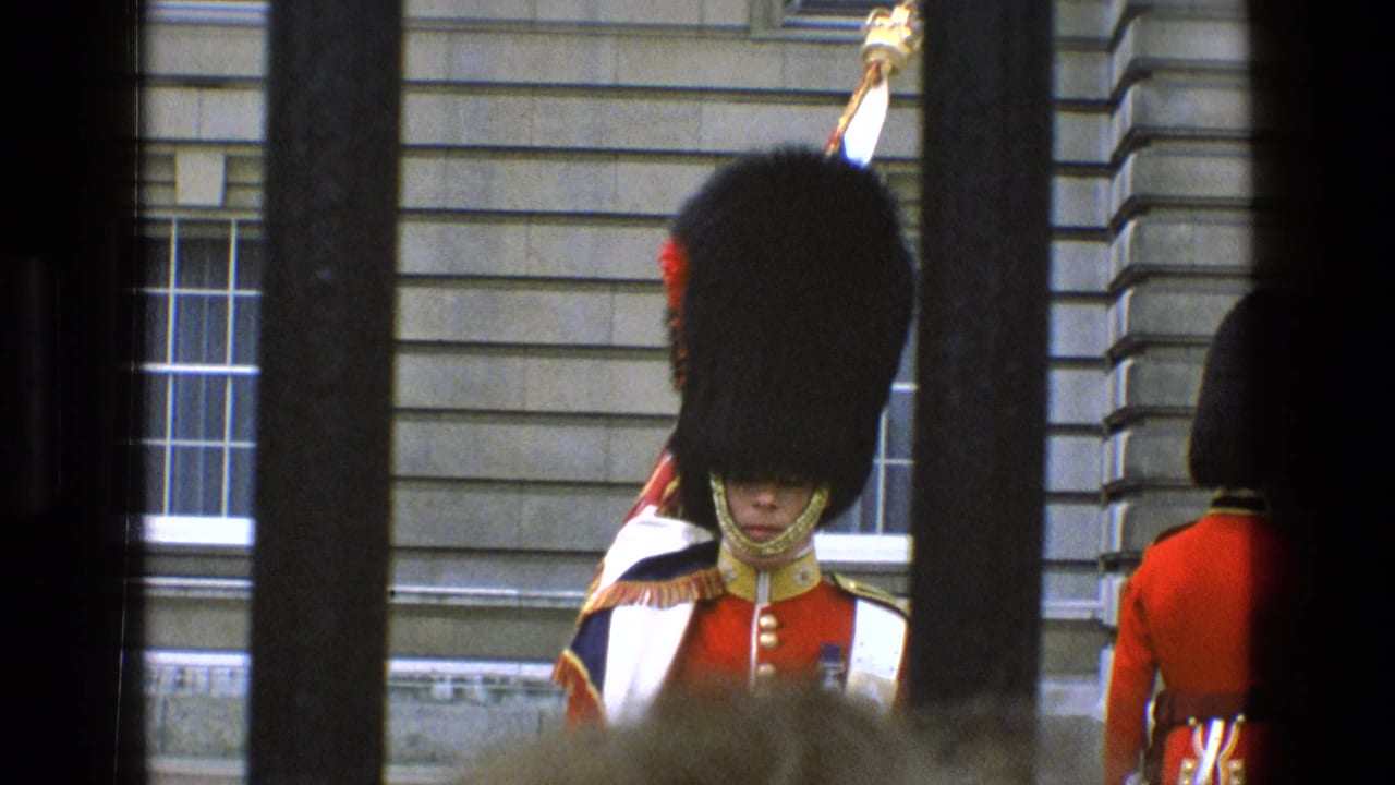 Guard in red tunic and bearskin hat stands near another guard by a stone building.