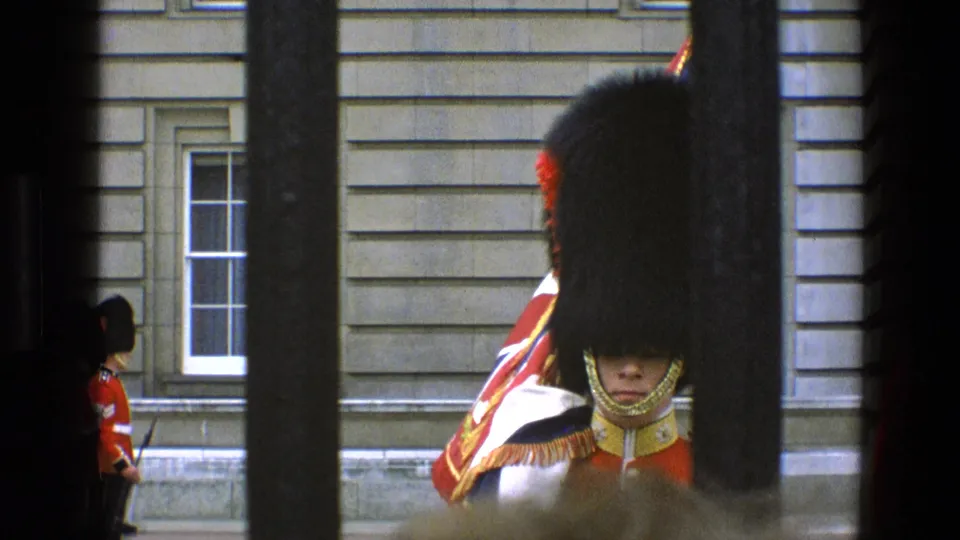 1969 England home-movie frame showing a close royal guard behind gate bars with a second guard visible to the left.