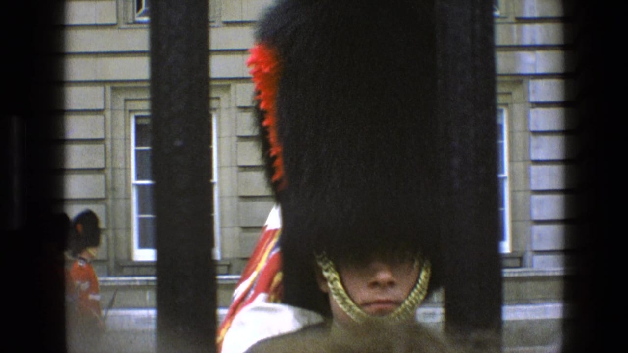 1969 England home-movie frame of a British guard in red ceremonial dress viewed through foreground bars and crowd obstruction.