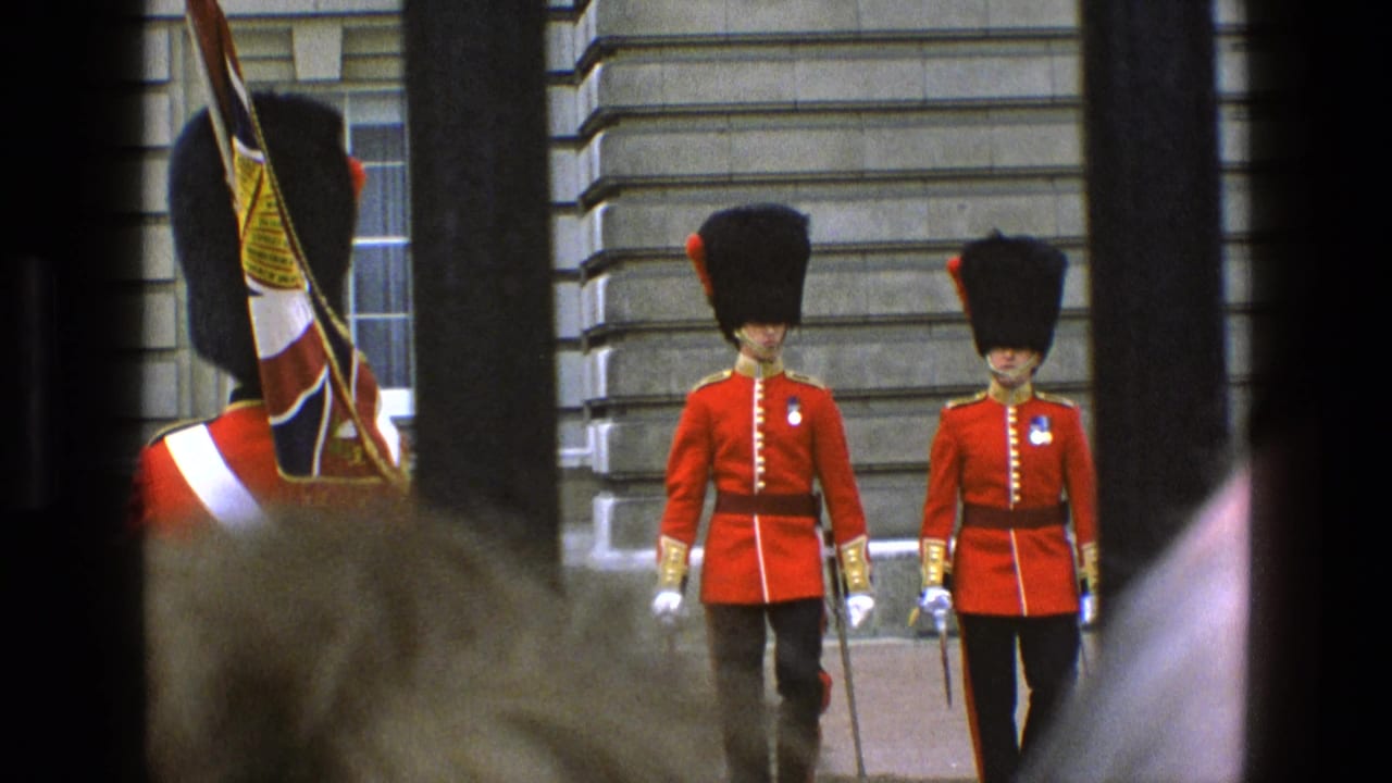 Two guards in red uniforms march near a stone building behind railings.