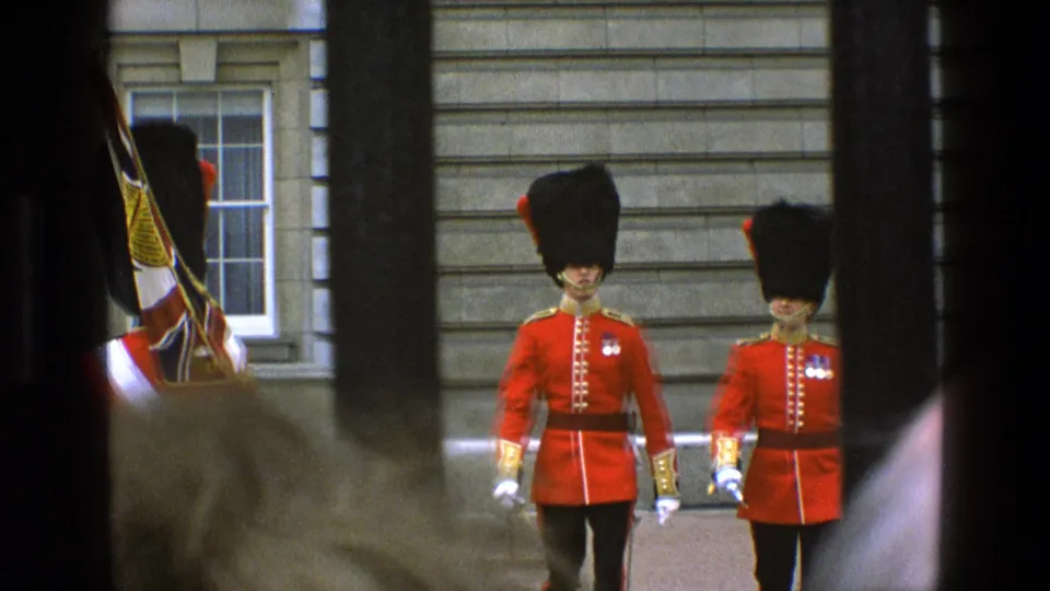 Two British guards in ceremonial uniform march through a barred viewpoint, with a Union Jack standard partly visible at left.