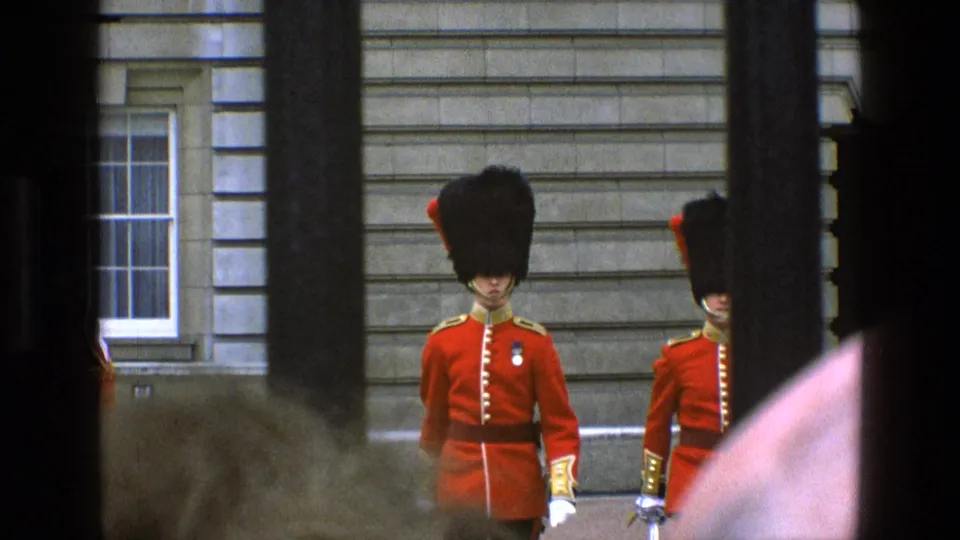 1969 England frame of British guards in red tunics and bearskin hats, partly obscured by railings and nearby spectators.
