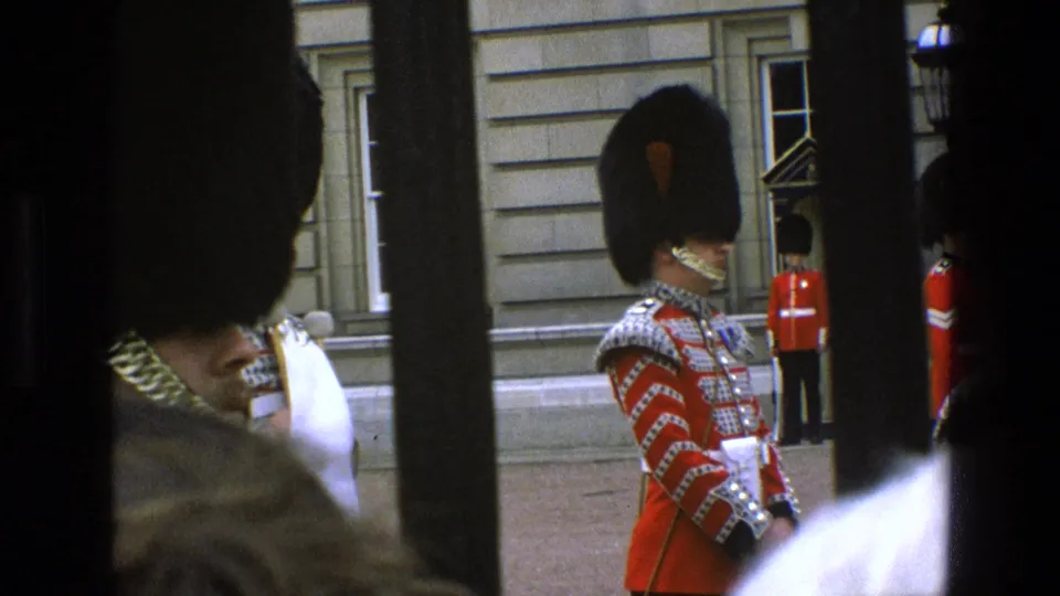 1969 England home-movie frame showing a guard in ornate ceremonial dress, partly blocked by dark bars and nearby spectators.