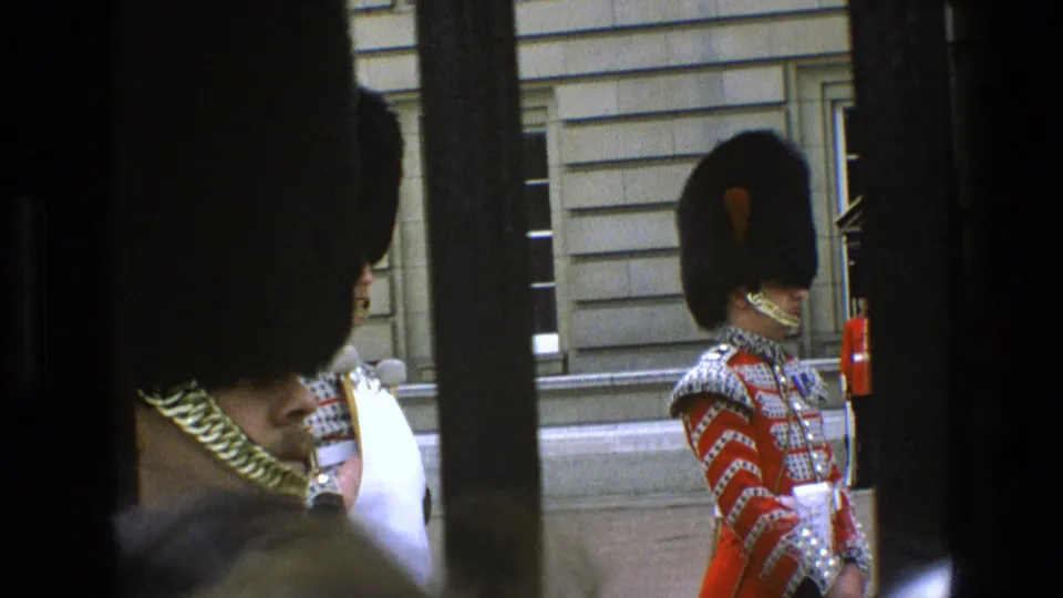 1969 England frame showing a side view of a ceremonial guard in detailed red uniform and bearskin hat, filmed through vertical bars.