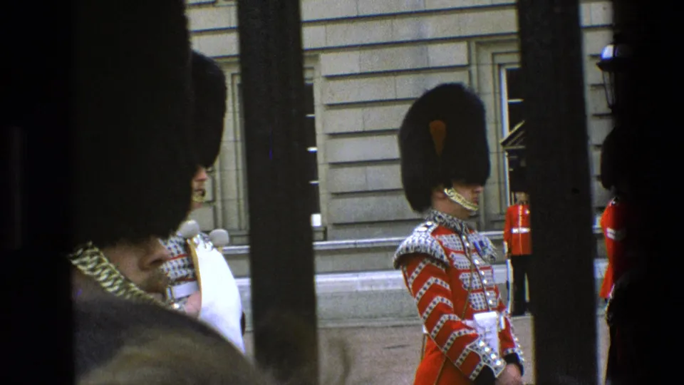 1969 England 8mm frame showing British ceremonial guards in red tunics and bearskin hats viewed through dark bars near a stone building.