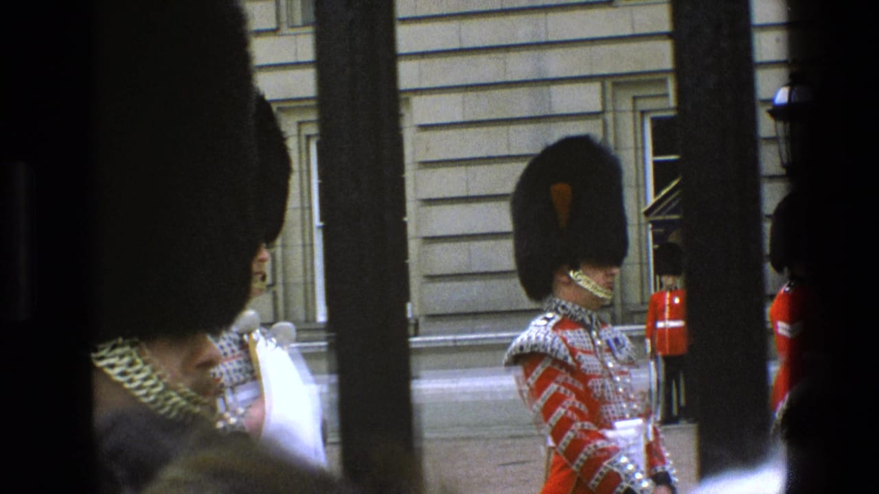 1969 England home-movie frame of a British ceremonial guard in red uniform and tall bearskin hat, partially blocked by vertical bars.