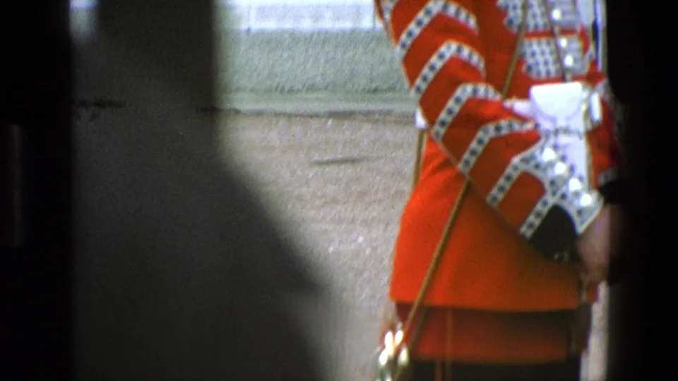 Close 8mm view of a ceremonial guard's red tunic, white belt, decorated cuff, and staff, partially obscured in England footage from 1969.
