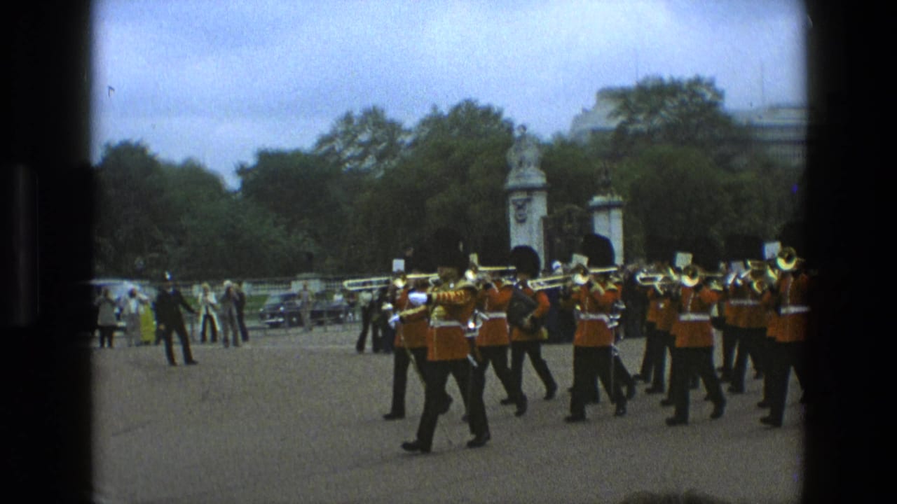 In 1969 England, guardsmen in red tunics and bearskin hats march with brass instruments across a paved parade ground.