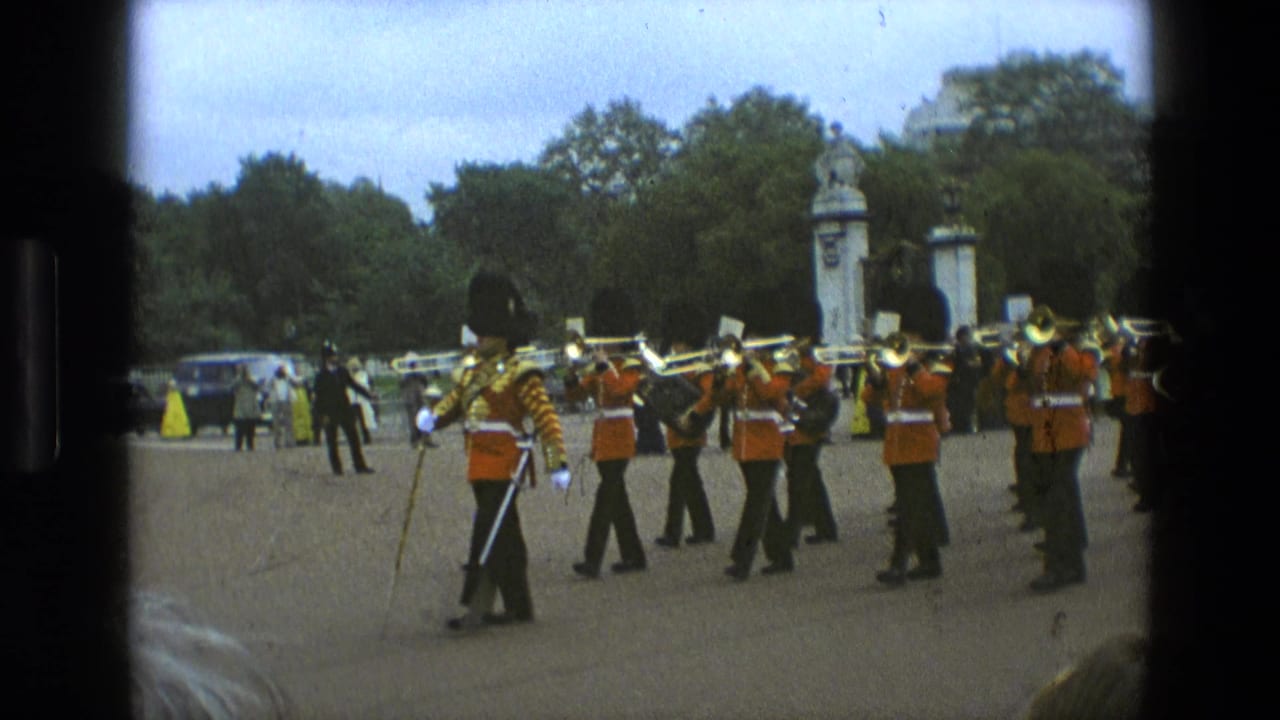 Drum major leading a line of brass players in red uniforms near stone gateposts and trees.