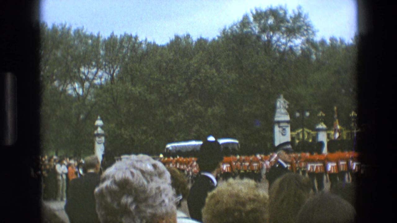 1969 England home-movie frame showing spectators watching Royal Guards in red coats near ornate gates, trees, and a stone monument.
