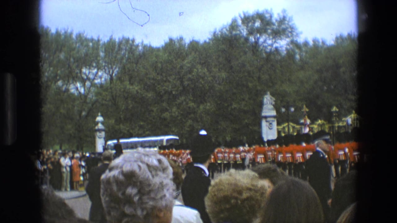 Royal Guards march in formation as spectators watch beside gates and trees.