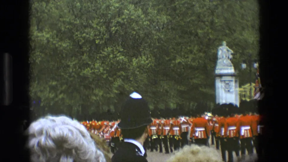 1969 England home-movie frame showing a police officer in a traditional helmet watching Royal Guards pass near a stone statue.