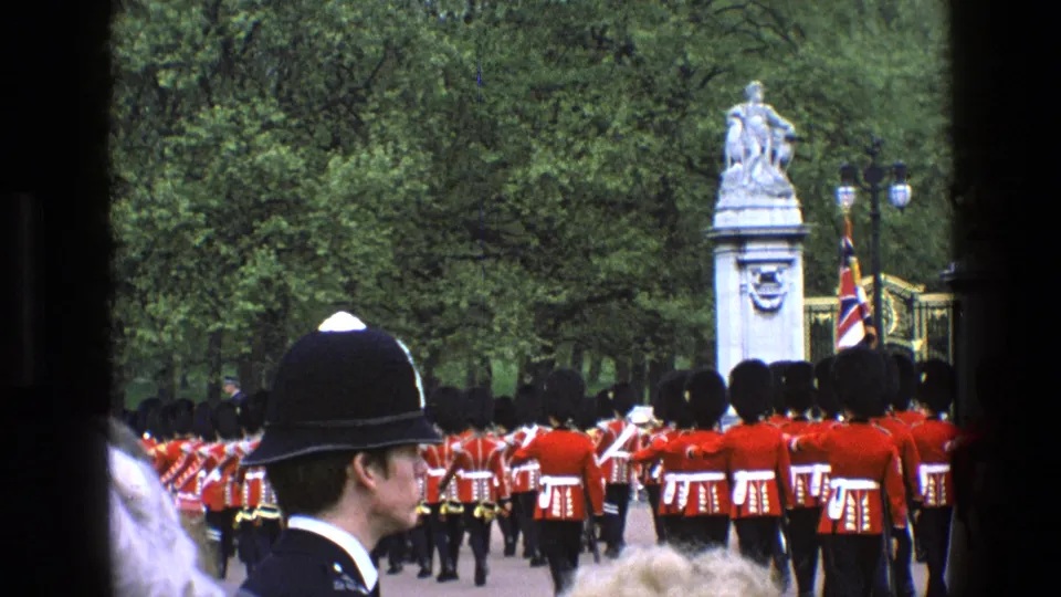 1969 England home-movie frame with a police officer in the foreground, Royal Guards in red coats, a flag, trees, and a stone statue.