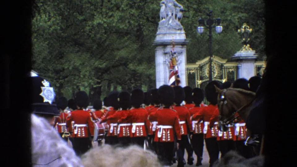 1969 England footage frame with guards in formation near ornate gates, a partial horse, and spectators visible at the edge.