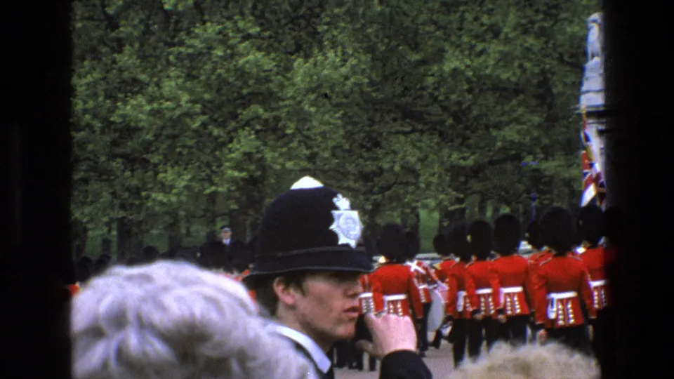 Crowd-level 1969 England home-movie view with a police officer in helmet watching ceremonial guards march away.