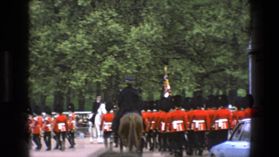 1969 England home-movie frame of guards in red tunics and bearskin hats marching near mounted personnel and spectators.