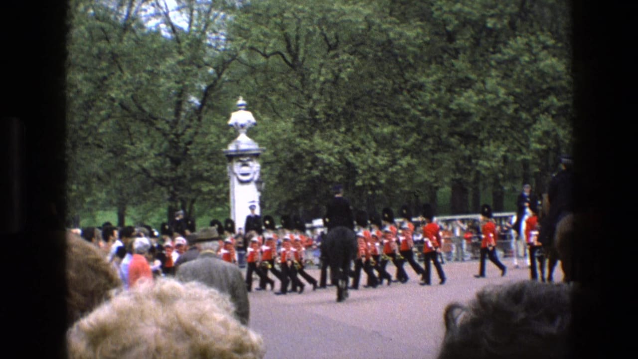 1969 England frame showing guards in red tunics crossing a park roadway during a ceremonial procession near Buckingham Palace.
