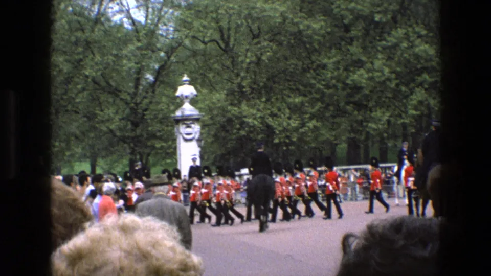 1969 England frame showing guards in red tunics crossing a park roadway during a ceremonial procession near Buckingham Palace.