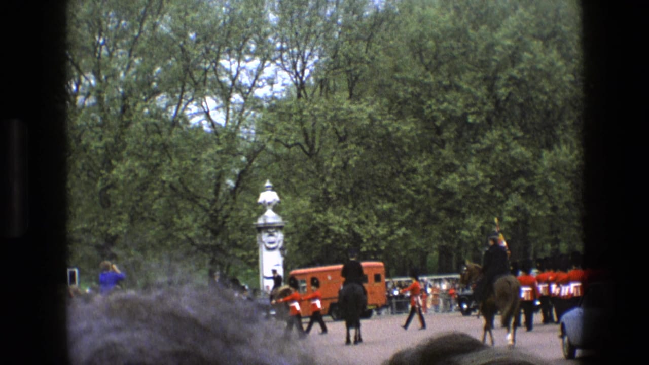1969 England home-movie view of guards in red tunics marching near a palace entrance, partly obscured by spectators in the foreground.