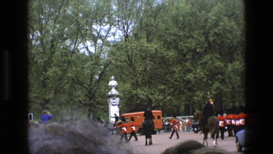 1969 England home-movie view of guards in red tunics marching near a palace entrance, partly obscured by spectators in the foreground.